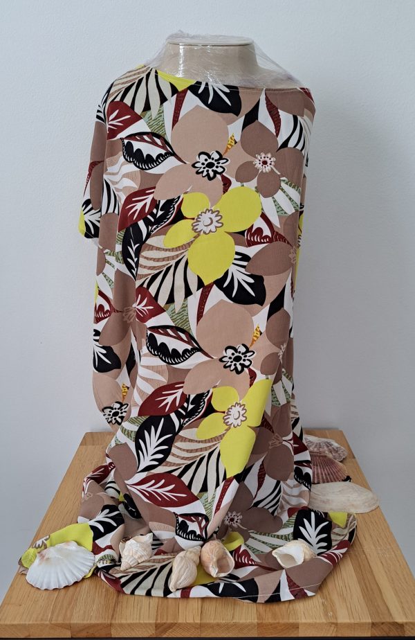 Robe de plage en viscose légère, imprimé fleurs et feuillages beiges, jaune et noirs, drapée sur un mannequin avec coquillages.