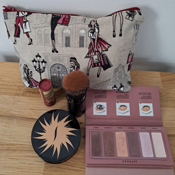 Trousse à maquillage “Shopping” en toile imprimée avec des motifs féminins représentant des silhouettes de femmes élégantes faisant du shopping, sur fond clair, avec une fermeture éclair dorée et intérieur doublé.