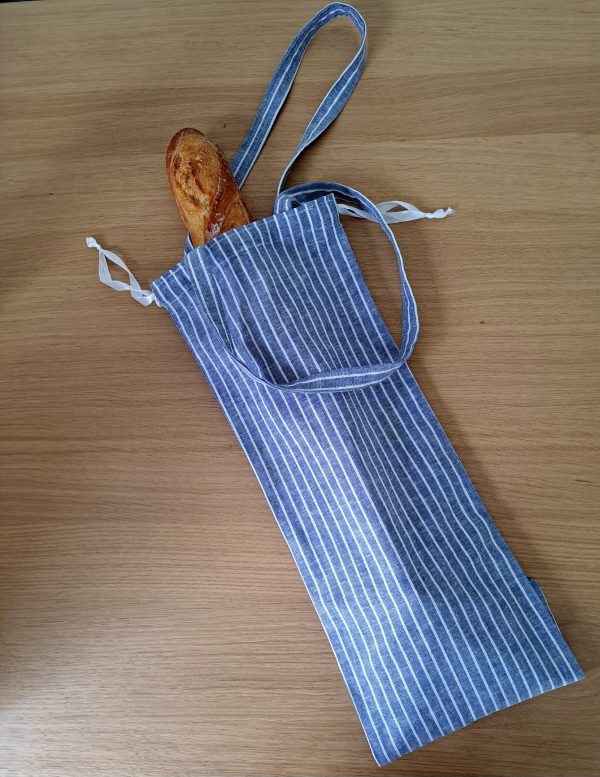 Sac à pain "Au Fil du Pain" en tissu rayé bleu et blanc, présenté debout et ouvert, avec un cordon de fermeture, posé sur une table en bois clair.