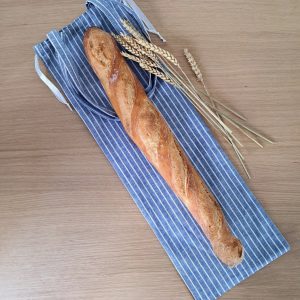 Sac à pain "Au Fil du Pain" en tissu bleu rayé, ouvert, laissant apparaître une baguette dorée posée sur une table en bois clair