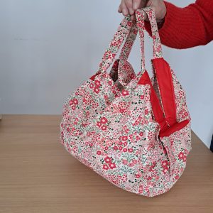Sac cabas Pli Vite modèle Capucine en coton artisanal avec motifs floraux rouges et blancs, porté à l'épaule dans une rue commerçante.