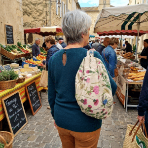 Personne portant un sac à motif floral Bocage dans un marché de village, marchant entre les étals de fruits, légumes, fromages et produits artisanaux, avec des bâtiments en pierre et une ambiance chaleureuse de marché local.