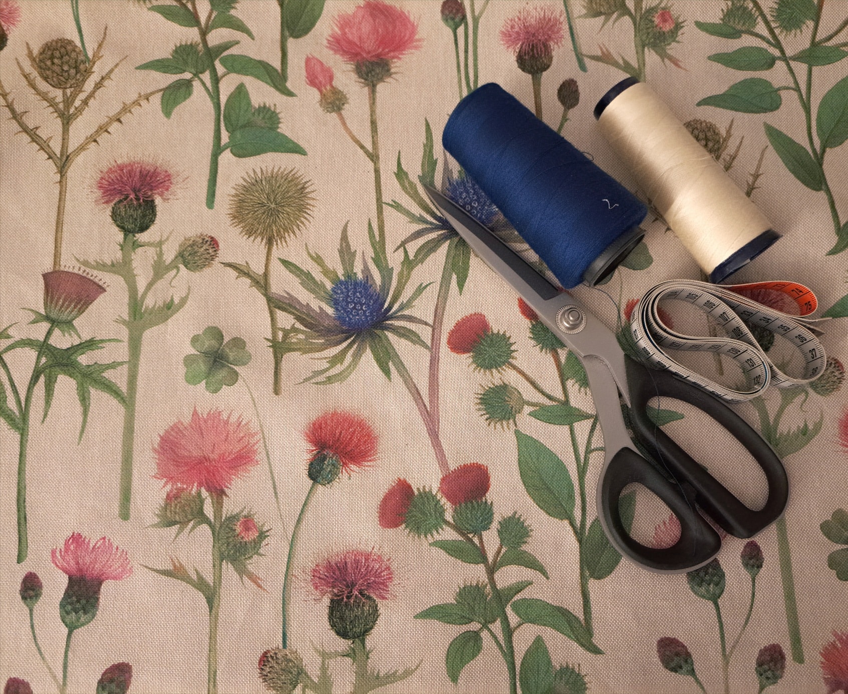 Outils de couture posés sur un tissu imprimé de fleurs sauvages et de chardons, comprenant une paire de ciseaux métalliques, deux bobines de fil bleu marine et écru, ainsi qu’un mètre ruban enroulé.