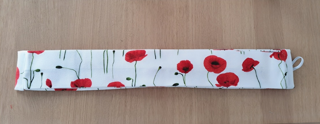 Tissu plié au motif de coquelicots rouges avec une boucle blanche, posé sur une surface en bois, montrant les détails du motif et de la finition