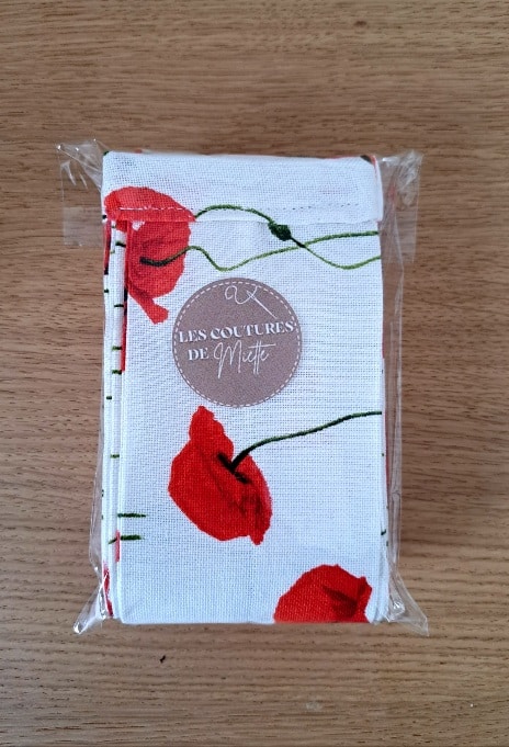 Pochette en tissu au motif de coquelicots rouges, emballée dans un sachet transparent avec une étiquette ronde “Les Coutures de Miette”, posée sur une surface en bois.